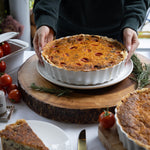 Person holding a dish of quiche with tomatoes and herbs on a wooden board.