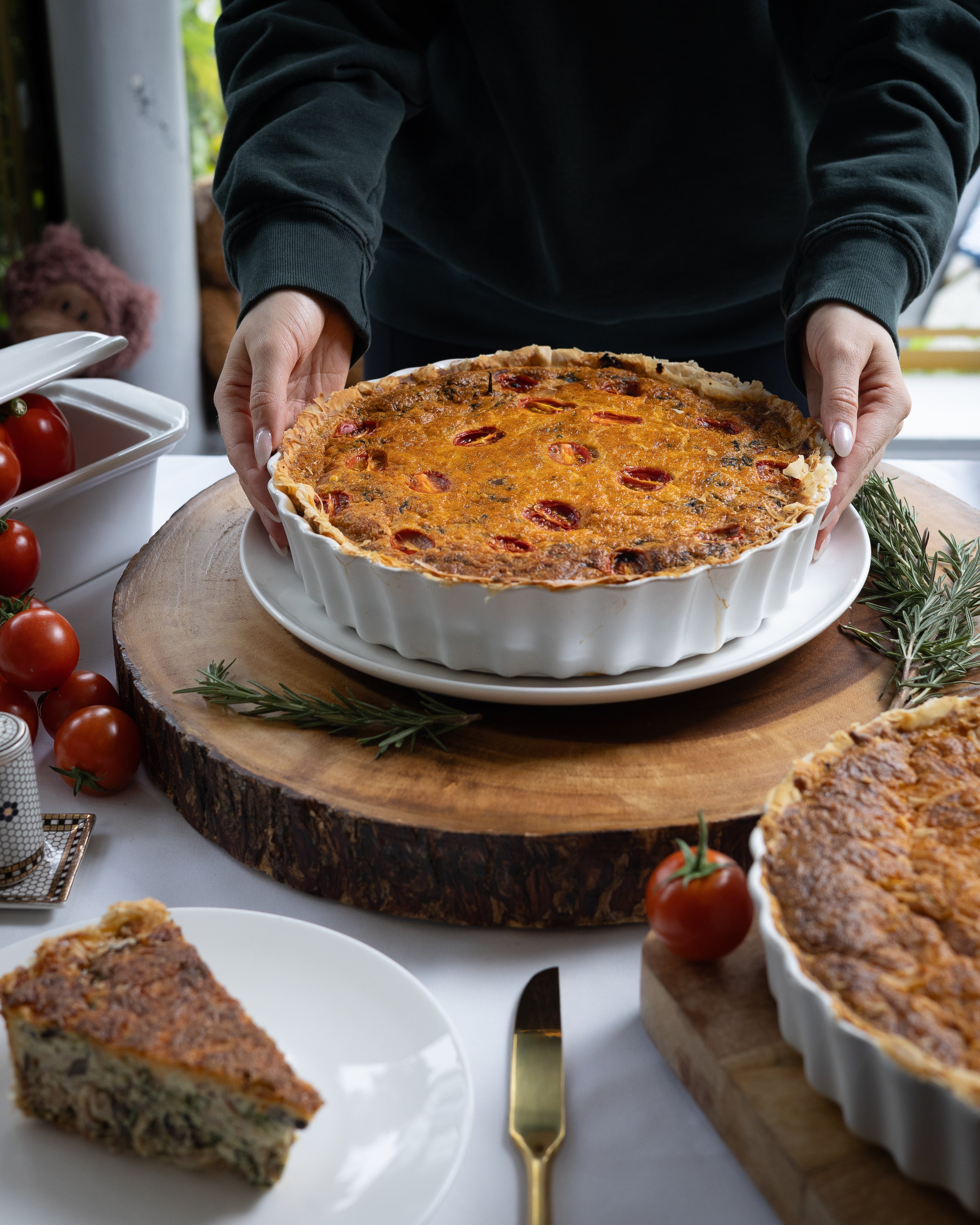 Person holding a dish of quiche with tomatoes and herbs on a wooden board.