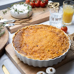 Quiche on a wooden cutting board with tomatoes, bread, and drinks in the background.