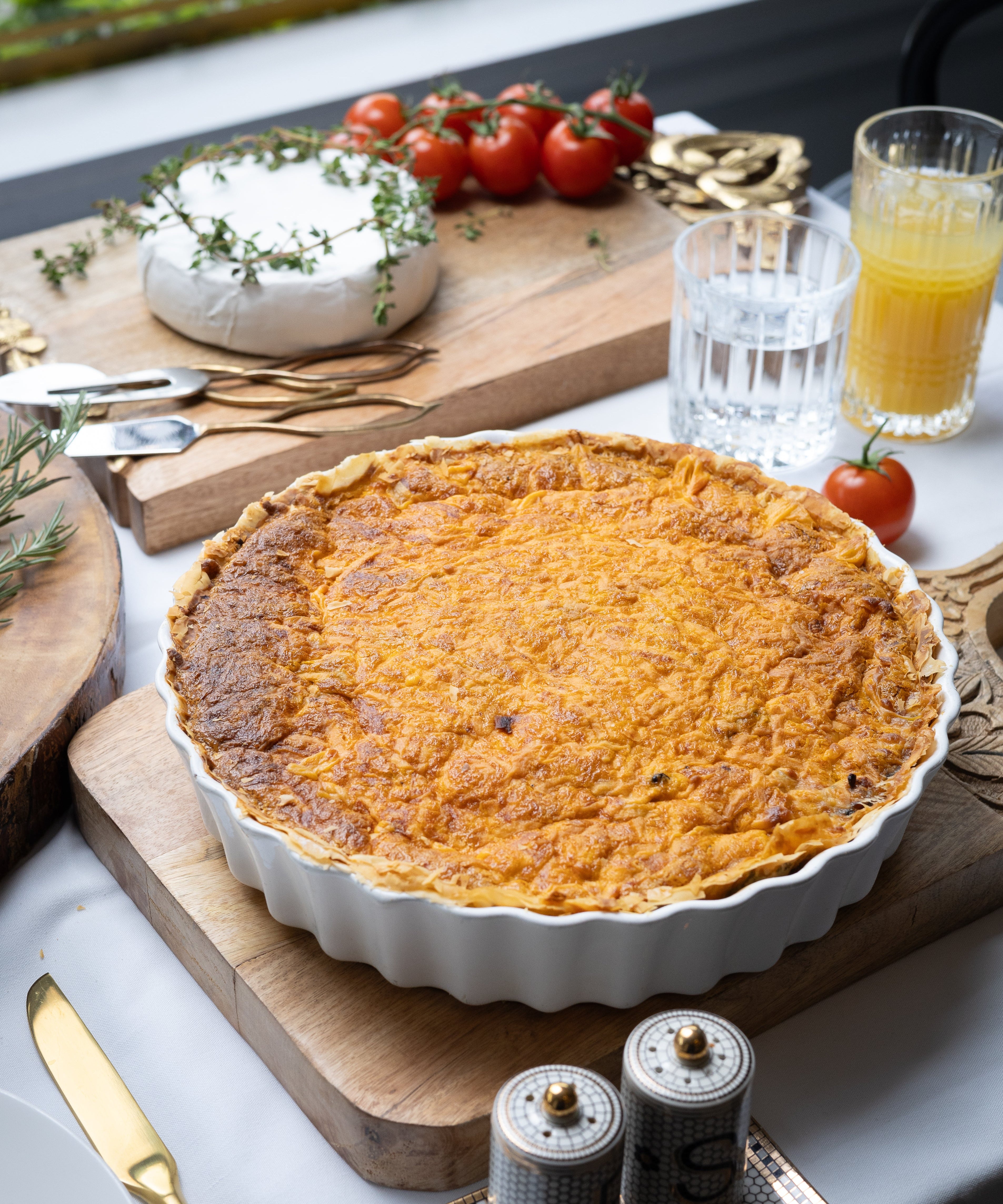 Quiche on a wooden cutting board with tomatoes, bread, and drinks in the background.