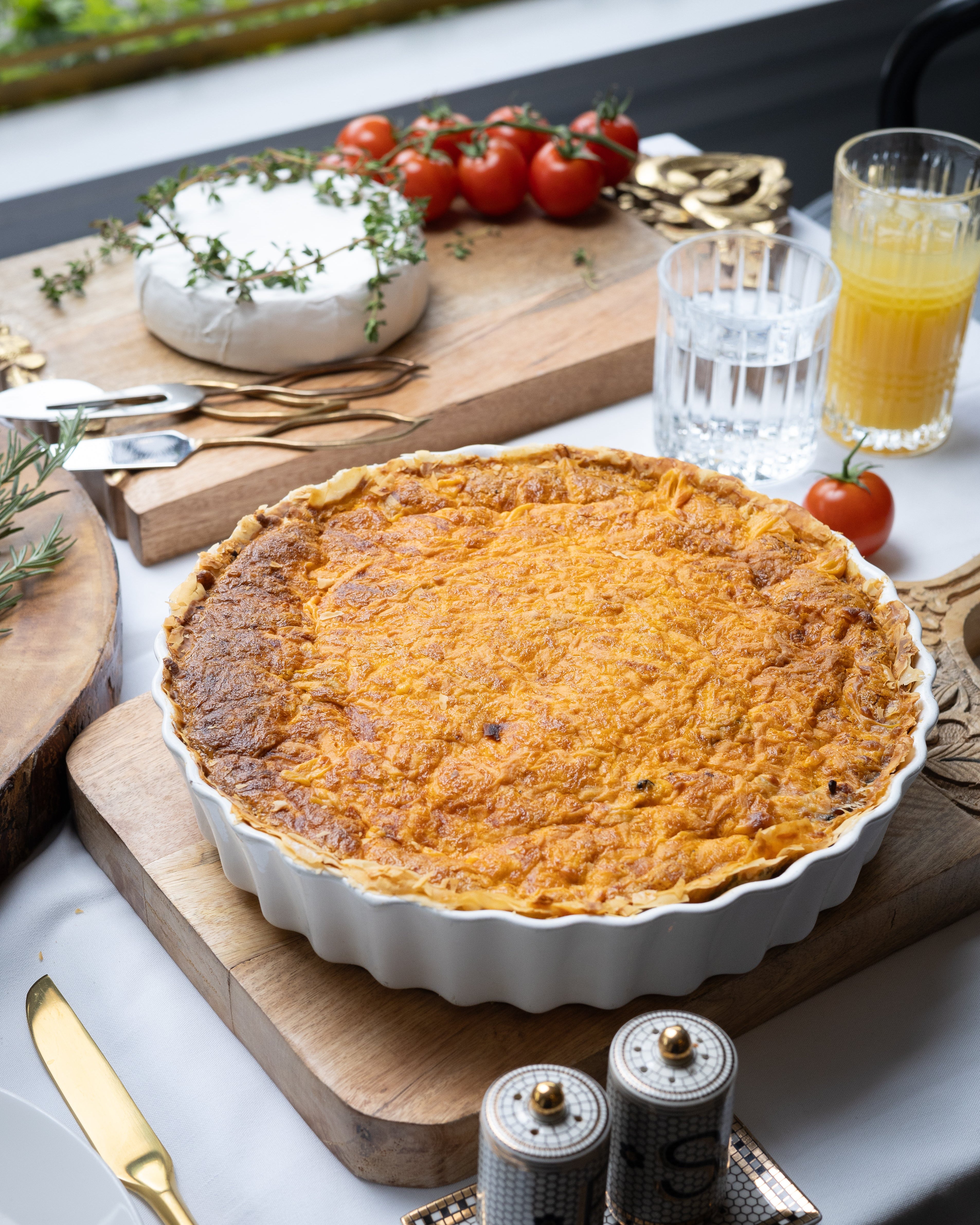 Quiche on a wooden cutting board with tomatoes, bread, and drinks in the background.