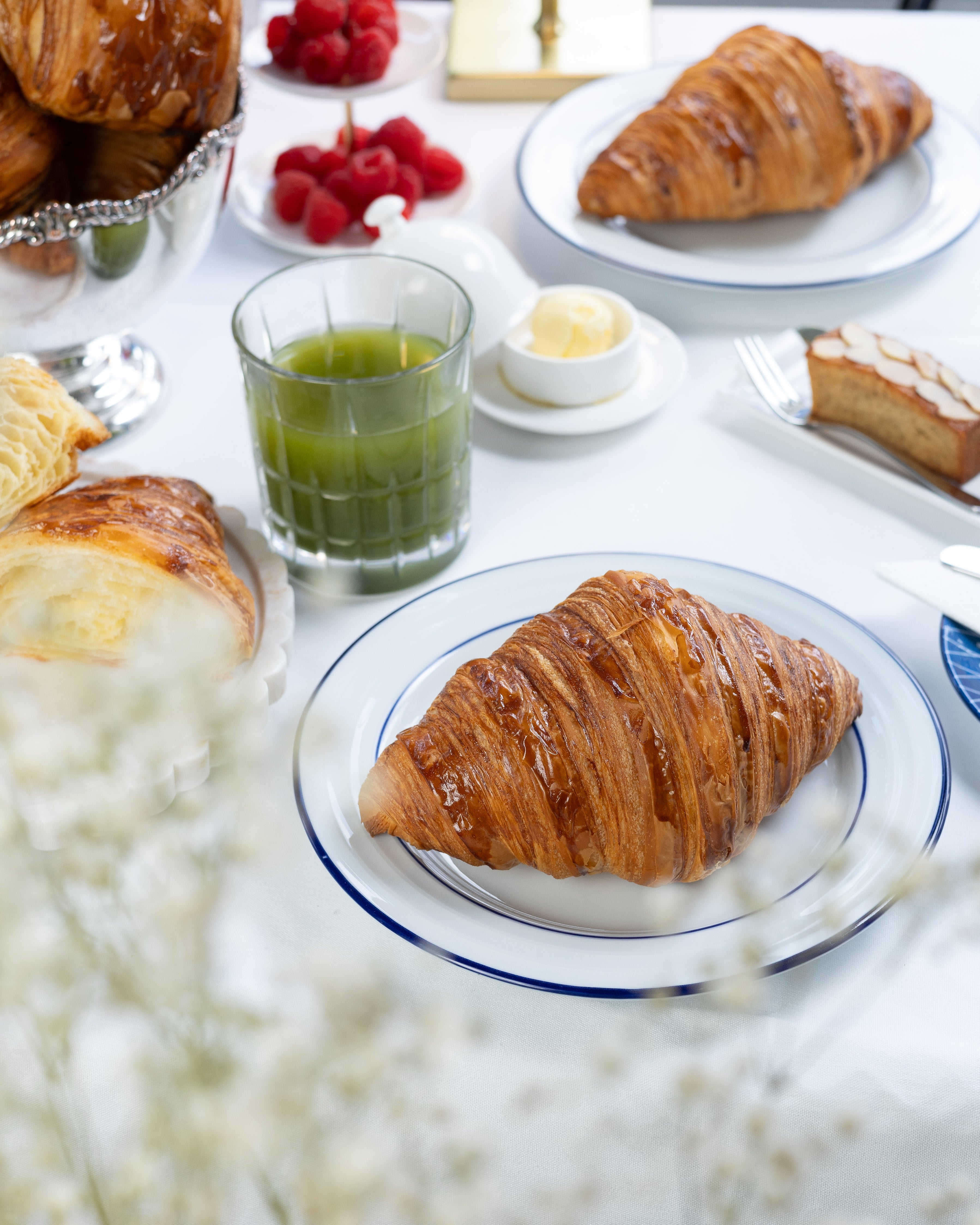 Table with croissants, raspberries, and a glass of green juice on a white tablecloth.