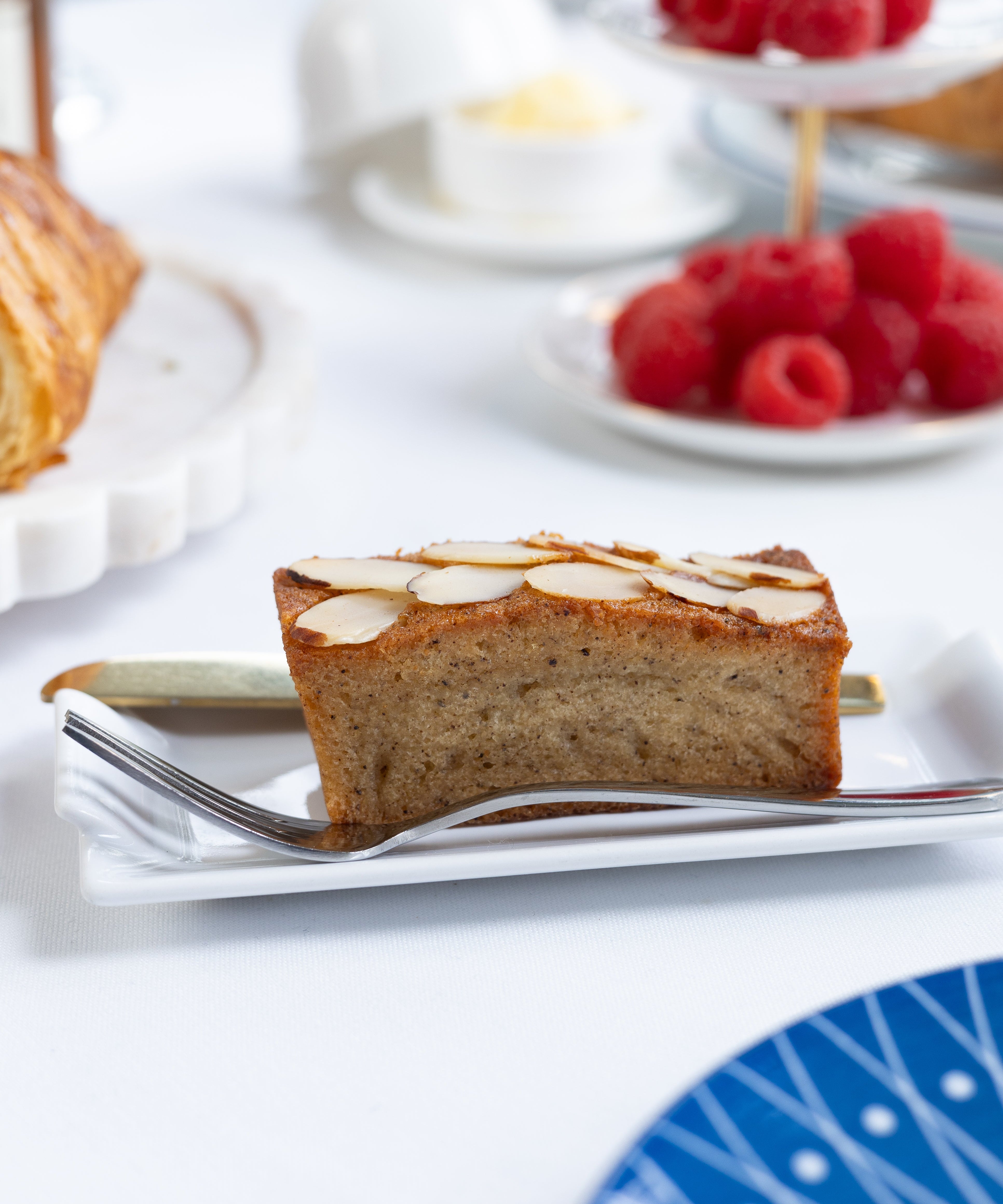 Slice of almond financier cake on a plate with raspberries and a teacup in the background
