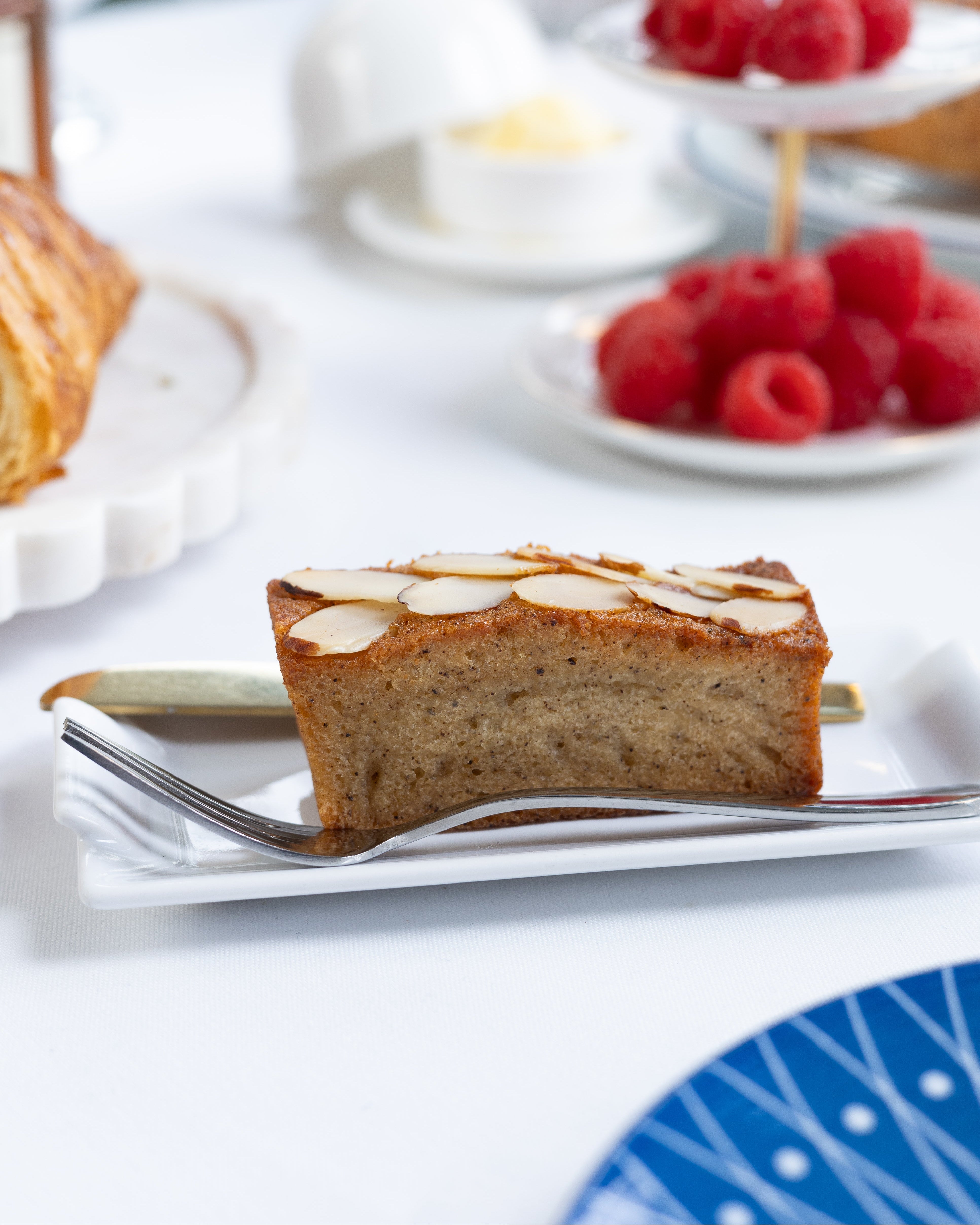 Slice of almond financier cake on a plate with raspberries and a teacup in the background