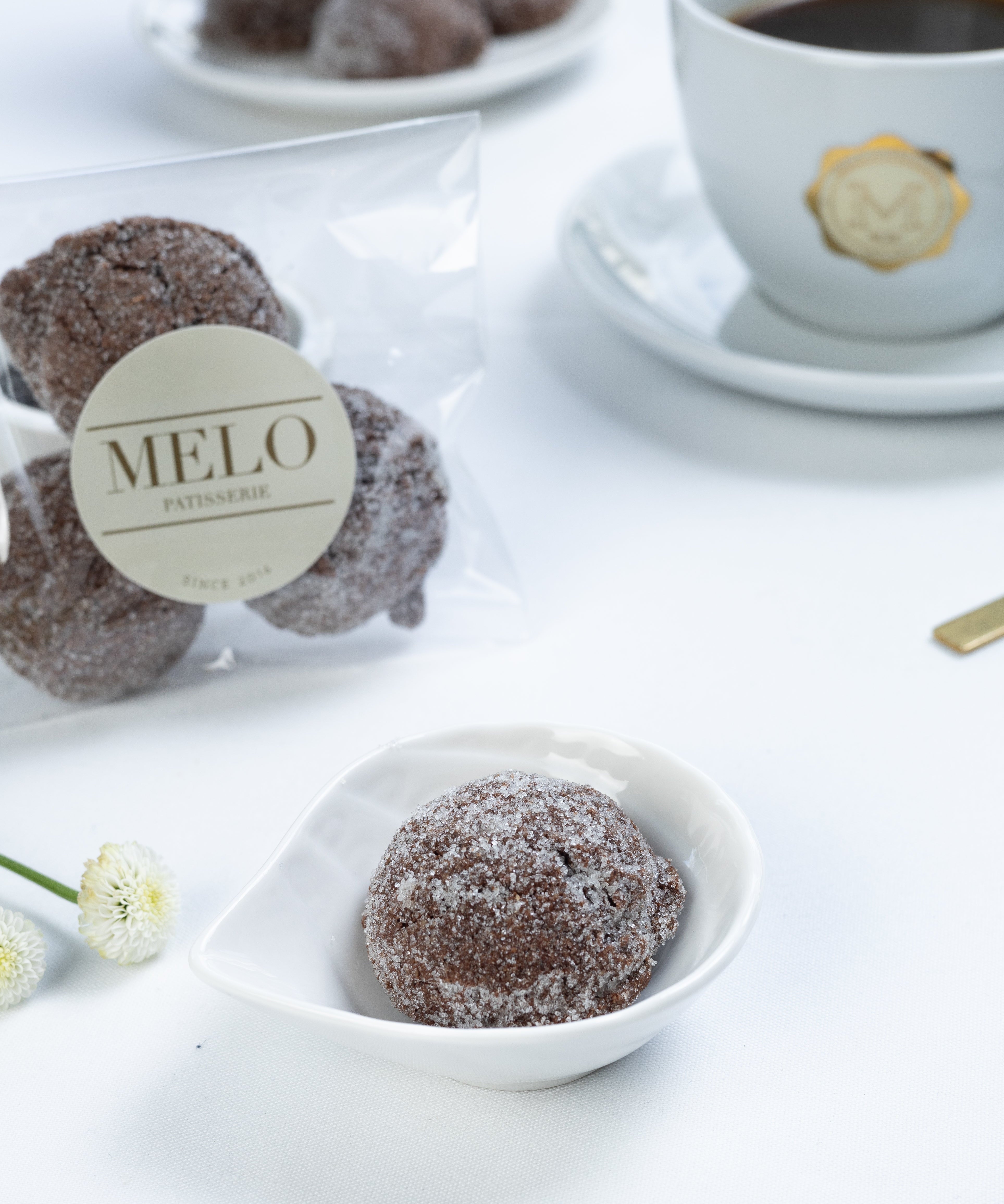 Almond Earl Grey cookies in a white dish with a cup of coffee and packaging labeled 'Melo' in the background.