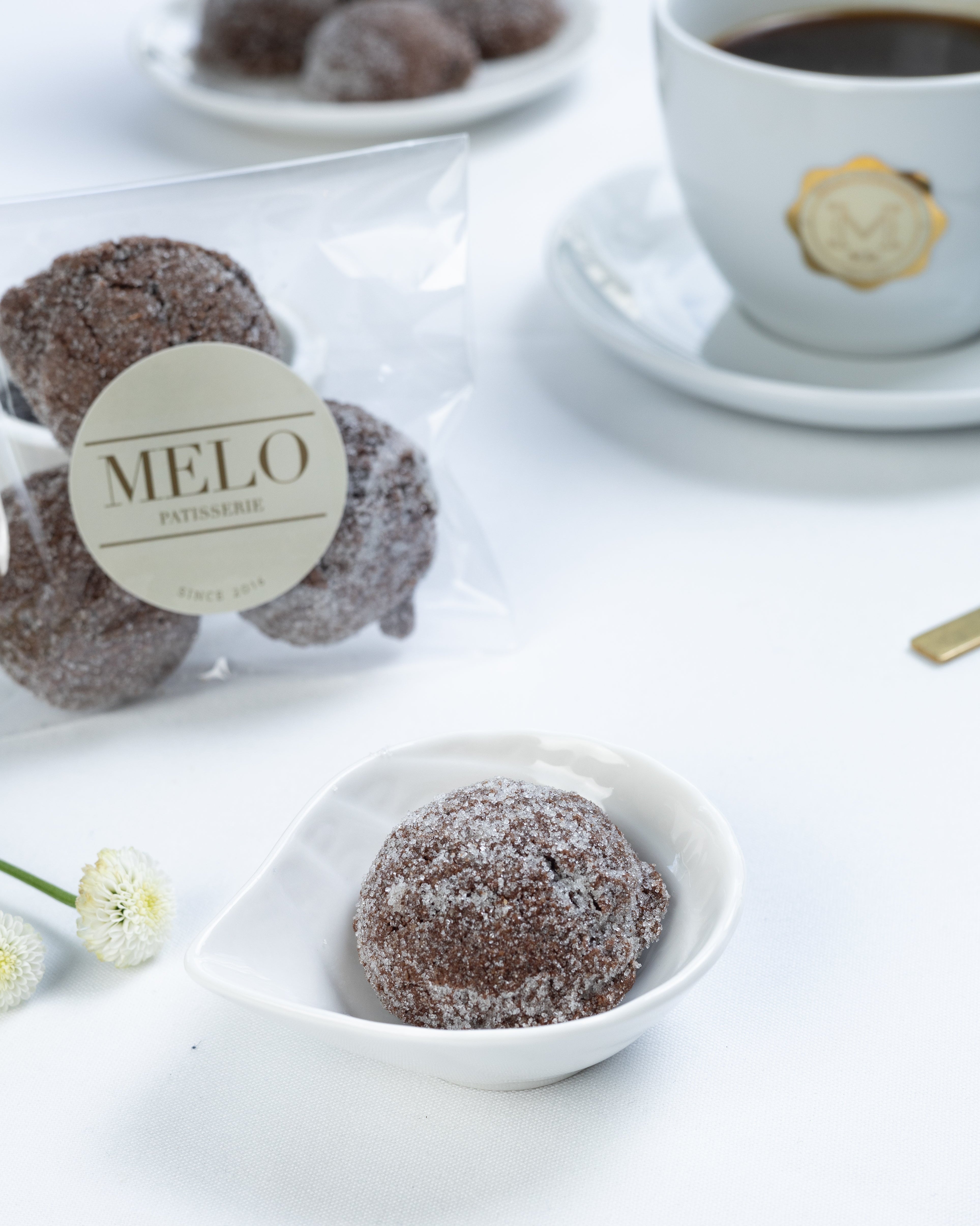 Almond Earl Grey cookies in a white dish with a cup of coffee and packaging labeled 'Melo' in the background.