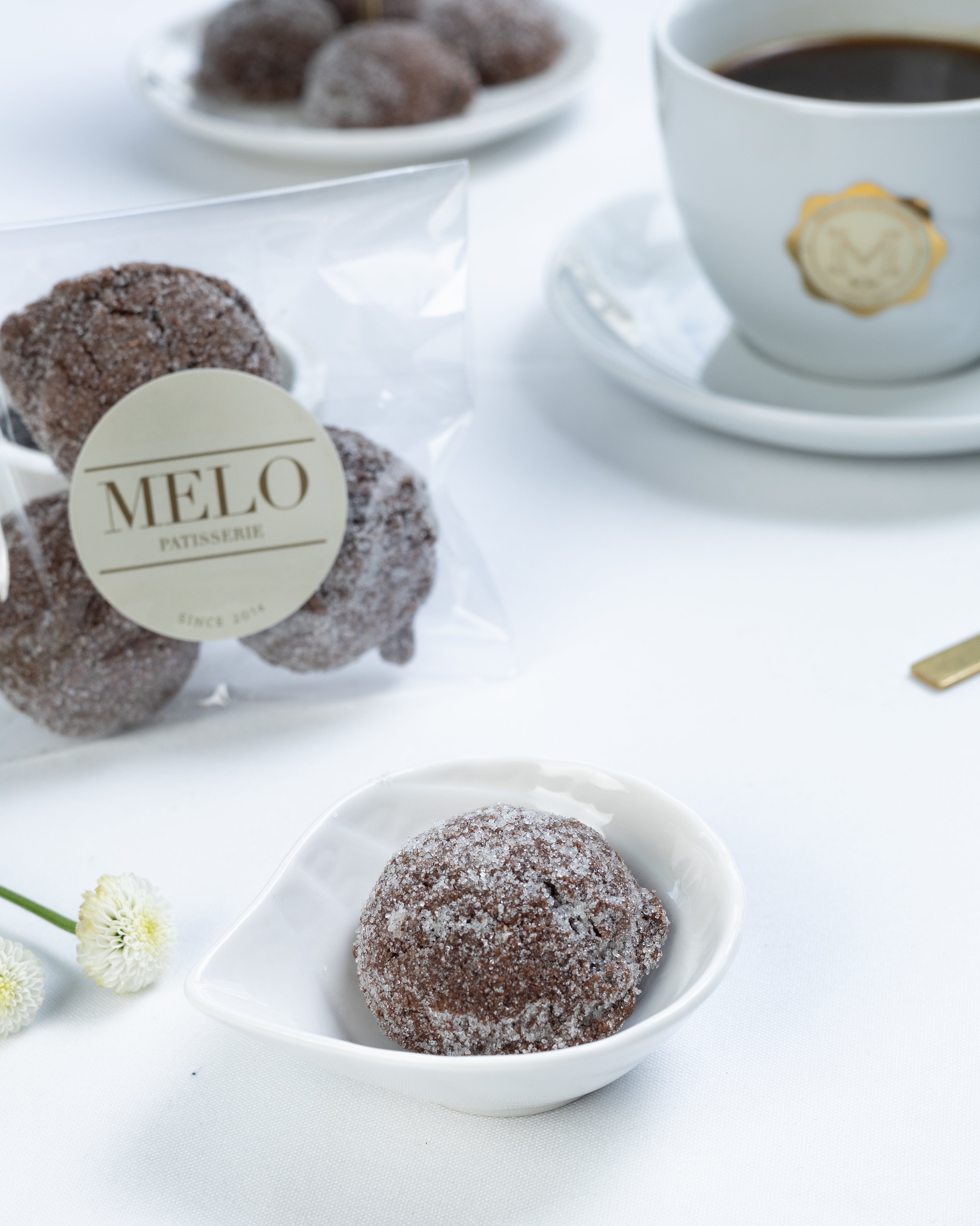 Almond earl grey cookies in a white dish with a cup of coffee and packaging labeled 'Melo' in the background.