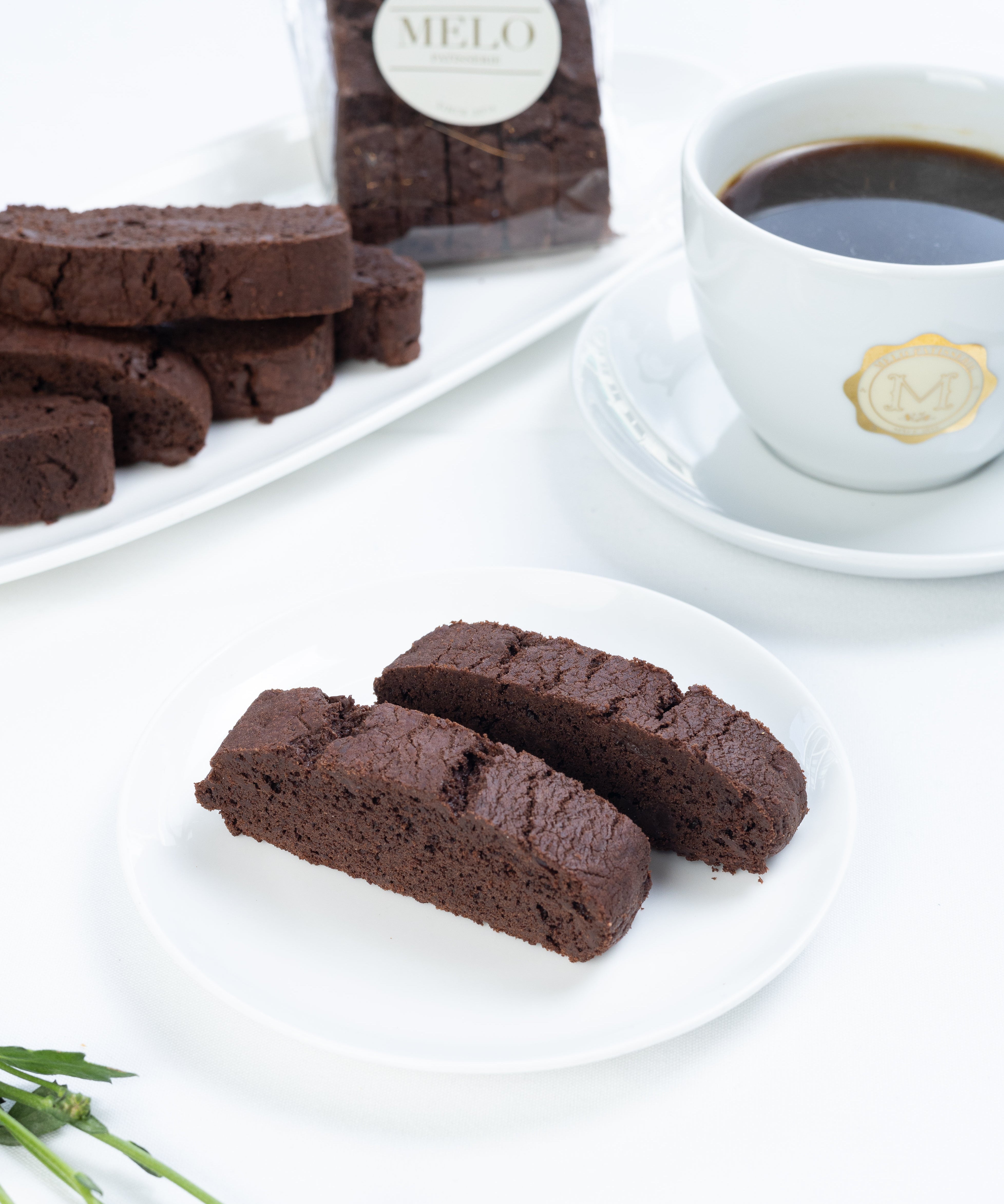 Chocolate biscotti on a white plate with a cup of coffee and flowers in the background.