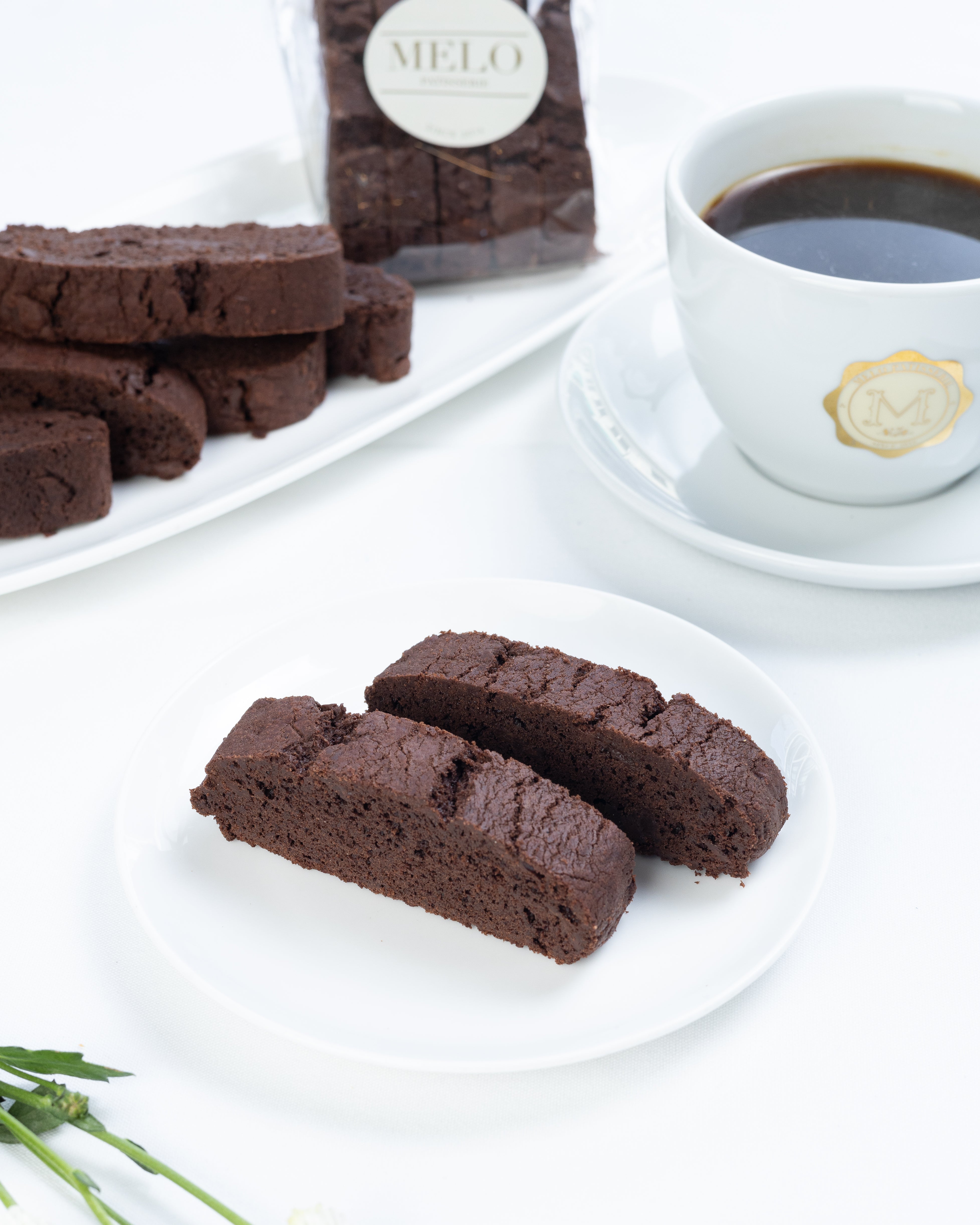 Chocolate biscotti on a white plate with a cup of coffee and flowers in the background.