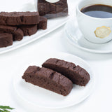 Chocolate biscotti on a white plate with a cup of coffee and flowers in the background.