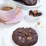 Chocolate cookie with nuts on a white plate with a cup of coffee and more cookies in the background.