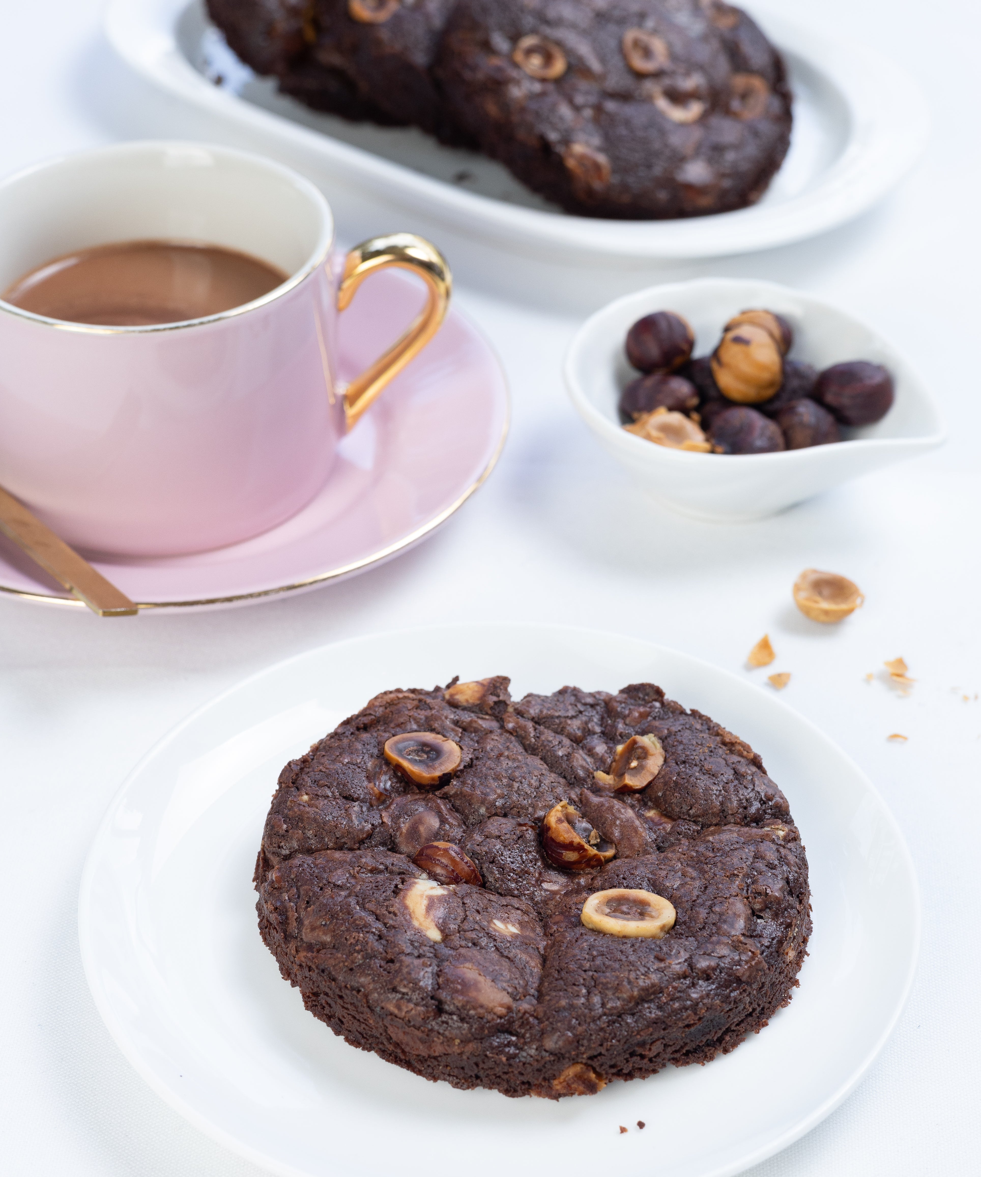 Chocolate cookie with nuts on a white plate with a cup of coffee and more cookies in the background.