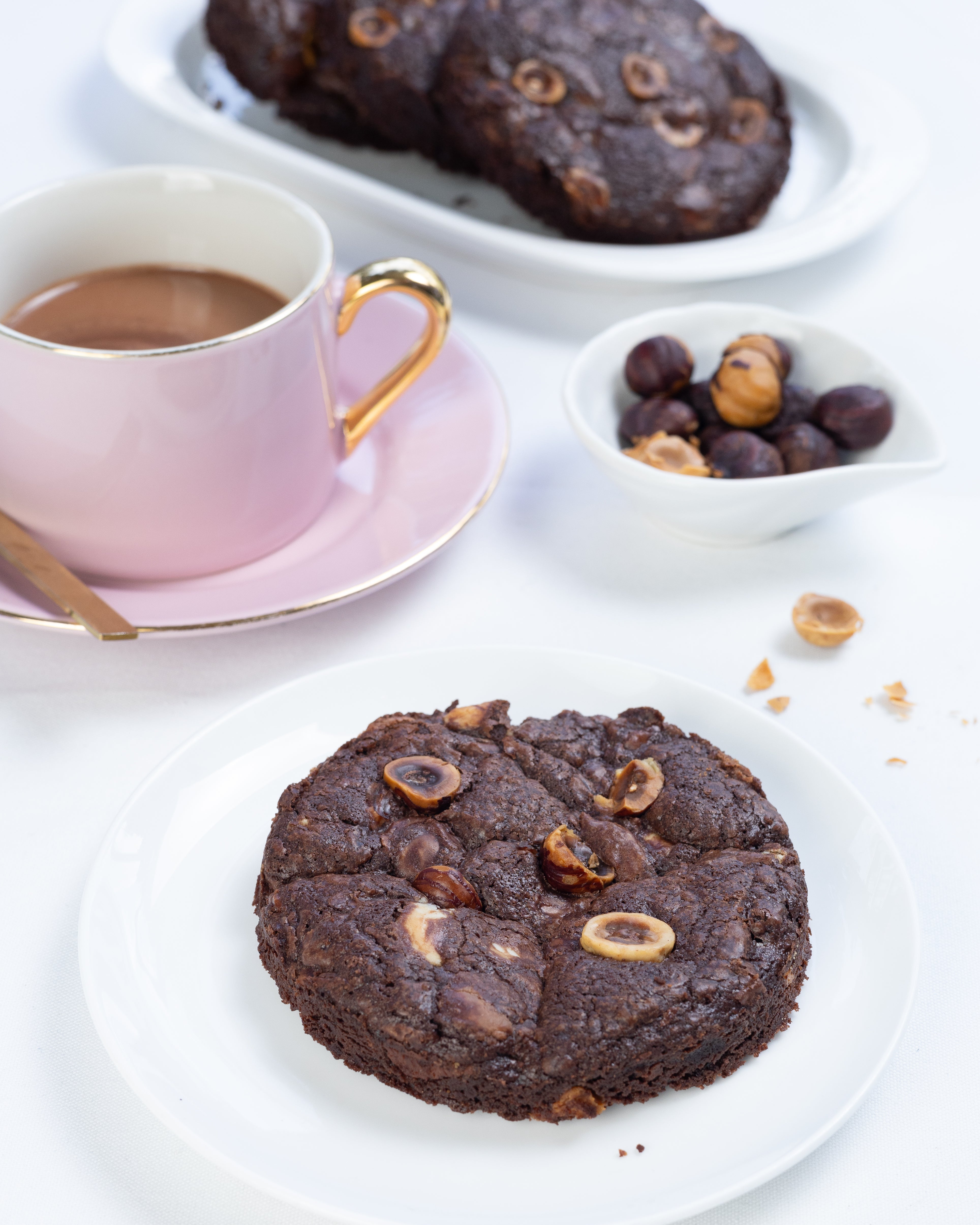 Chocolate cookie with nuts on a white plate with a cup of coffee and more cookies in the background.