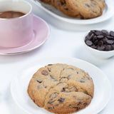 Chocolate chip cookies on a white plate with a cup of coffee and additional cookies in the background.