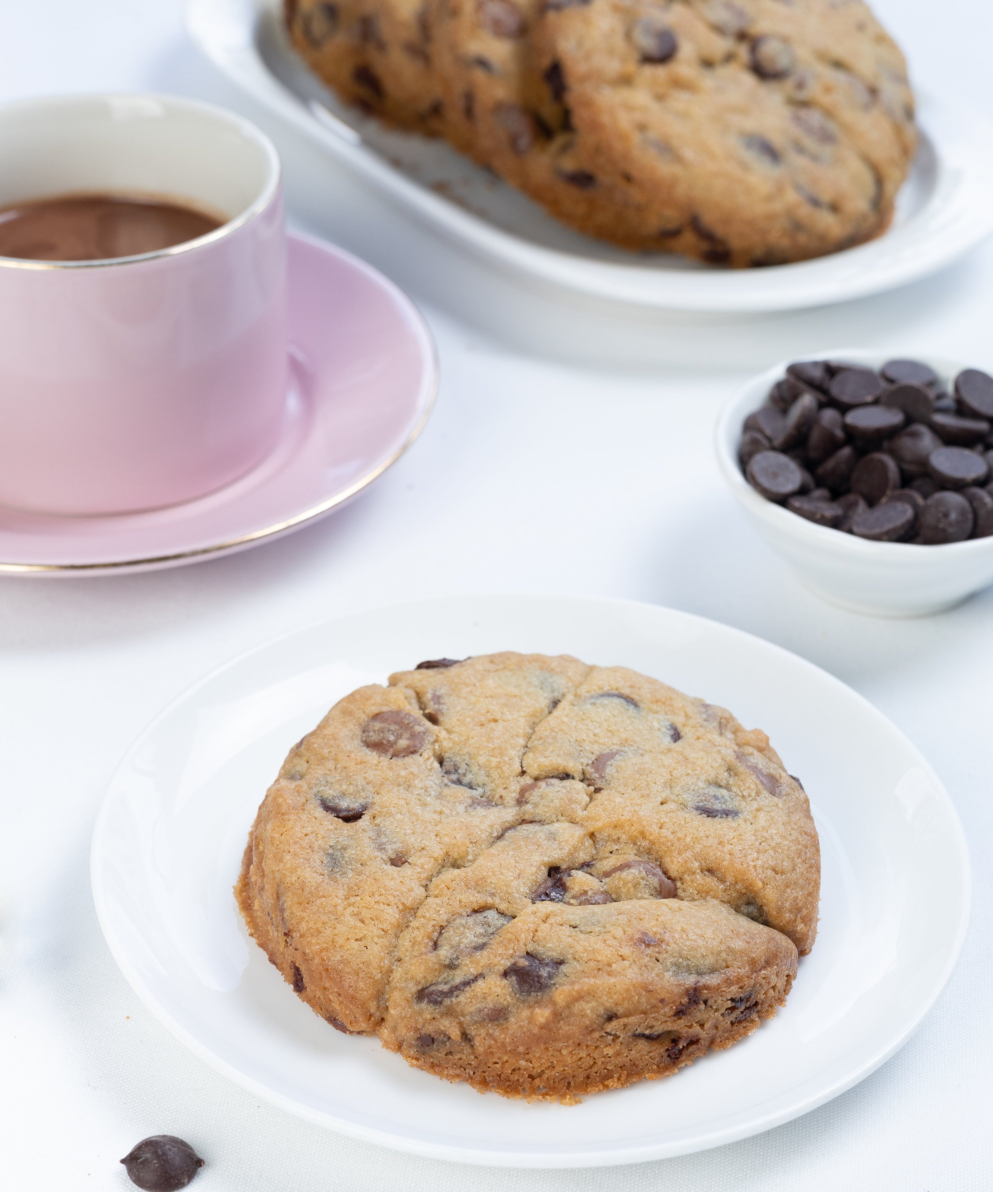 Chocolate chip cookies on a white plate with a cup of coffee and additional cookies in the background.