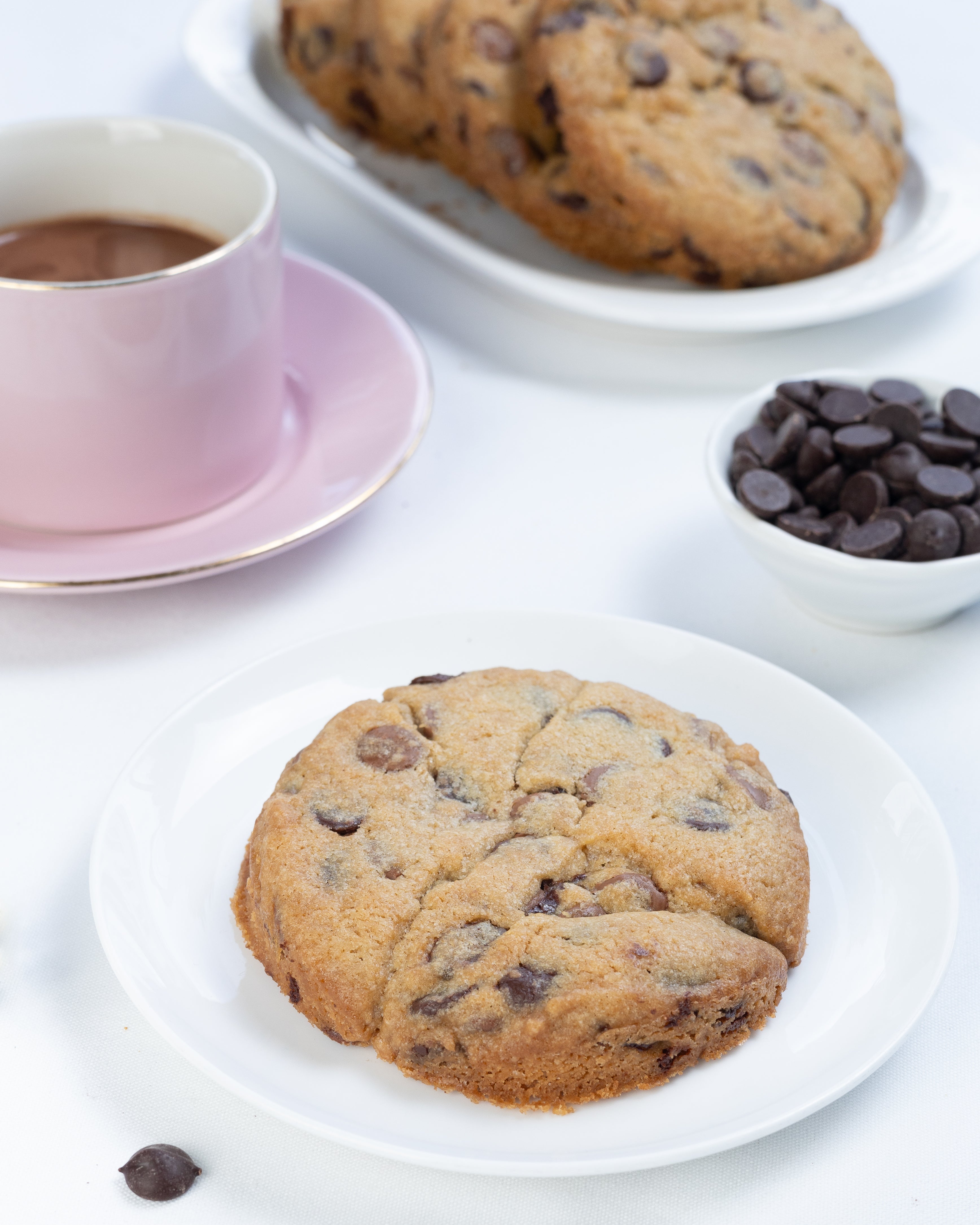 Chocolate chip cookies on a white plate with a cup of coffee and additional cookies in the background.