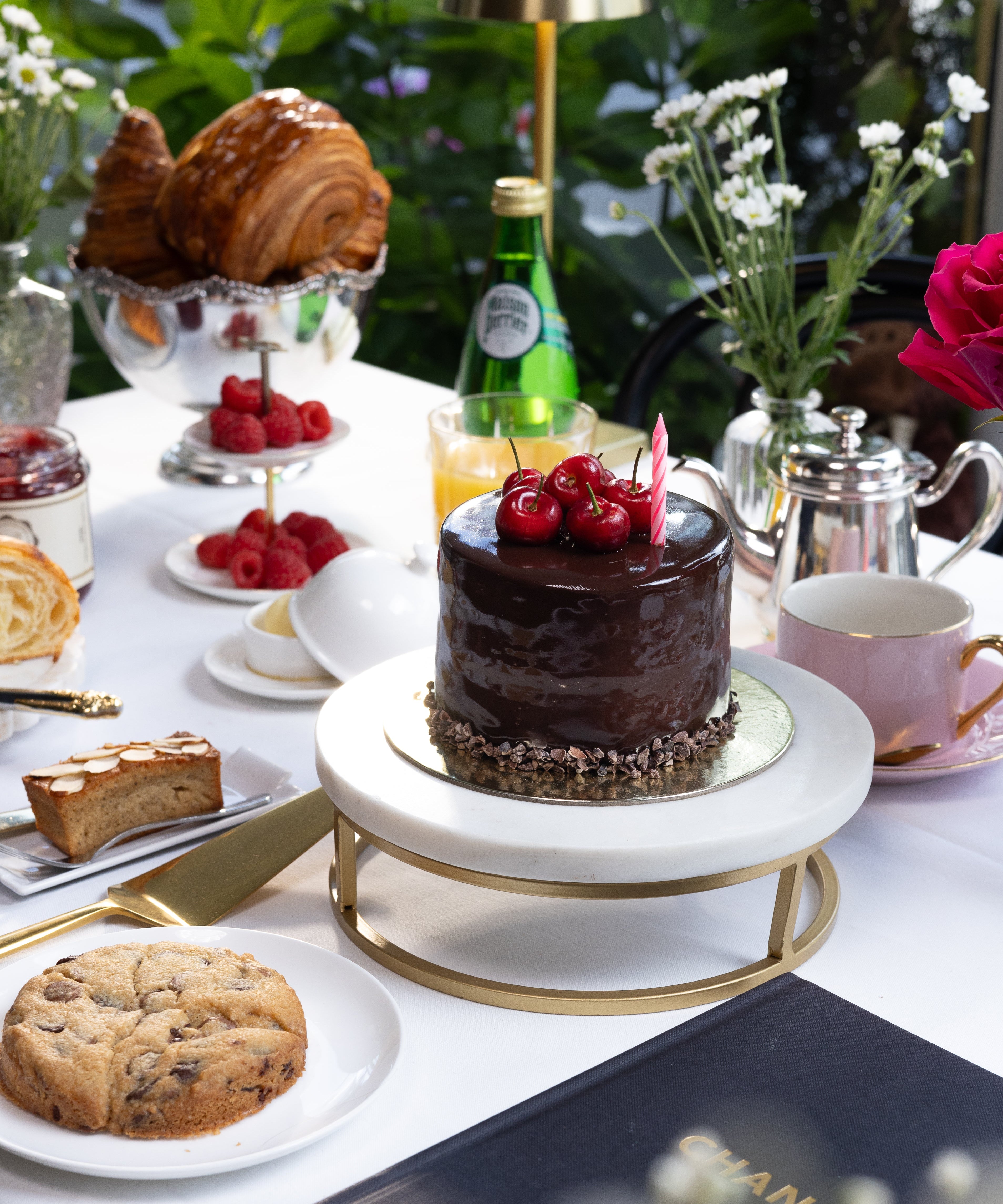 Table setting with a chocolate cake, cookies, and a Chanel menu in an outdoor setting.