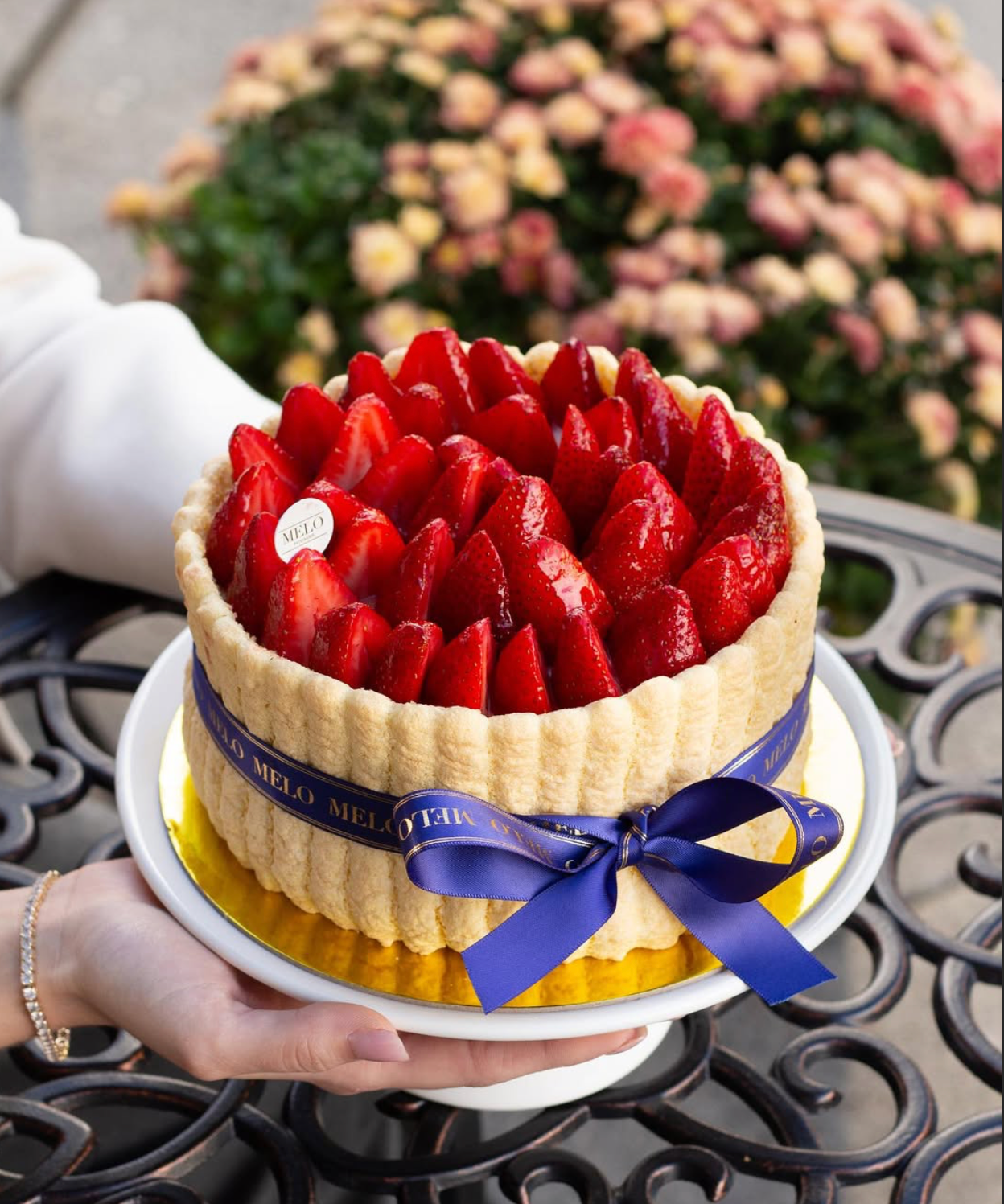 Cake with strawberries and a blue ribbon held by a person, with flowers in the background.