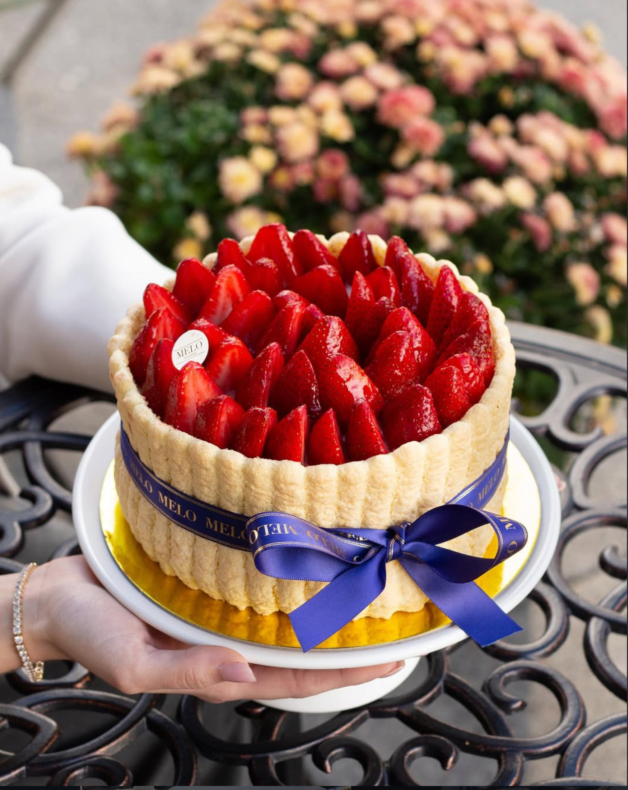 Cake with strawberries and a blue ribbon held by a person, with flowers in the background.